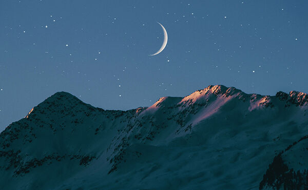 Crescent moon and stars shining over snow-covered mountain peaks at twilight.